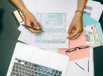 pexels-photo-8927687-8927687 Hands writing on tax documents with laptop, glasses, and currency on desk.